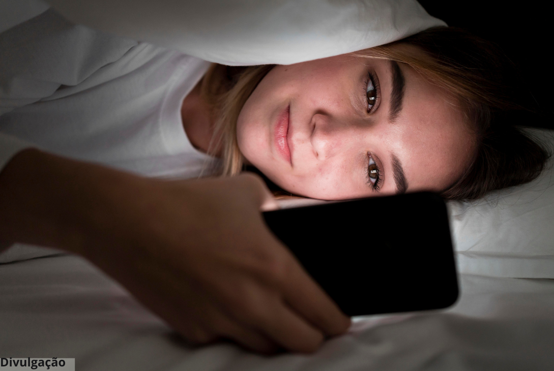 Foto ilustrativa de uma mulher embaixo de uma coberta na cama e mexendo no celular.