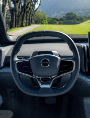 A imagem mostra o interior de um carro da Volvo, com destaque para o volante com o logo da marca e uma tela central de grandes proporções que exibe informações como GPS, reforçando a ideia de tecnologia, sofisticação e conectividade embarcada no veículo.