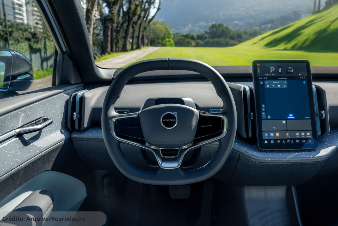 A imagem mostra o interior de um carro da Volvo, com destaque para o volante com o logo da marca e uma tela central de grandes proporções que exibe informações como GPS, reforçando a ideia de tecnologia, sofisticação e conectividade embarcada no veículo.