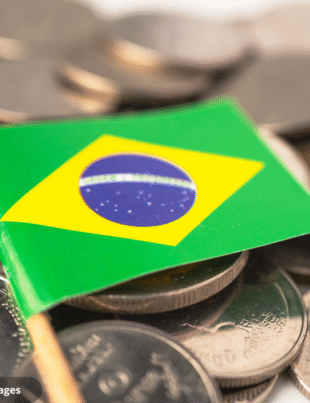 Imagem ilustrativa de um enfeite de bandeira do Brasil em cima de várias moedas nacionais.