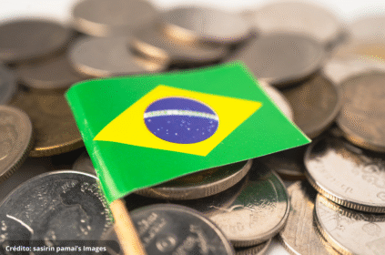 Imagem ilustrativa de um enfeite de bandeira do Brasil em cima de várias moedas nacionais.