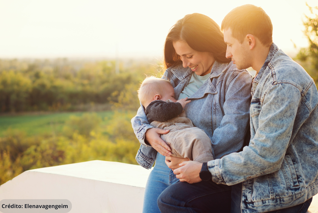 Foto ilustrativa de um momento afetuoso e íntimo entre uma família: uma mulher amamenta seu bebê enquanto o homem a acompanha carinhosamente, formando um abraço coletivo ao redor da criança. Esse retrato reflete não apenas a importância da amamentação, mas também o papel essencial da licença paternidade no fortalecimento do vínculo familiar. A presença ativa do pai desde os primeiros dias do bebê contribui para uma rede de apoio emocional e prática à mãe, além de favorecer o desenvolvimento saudável da criança.