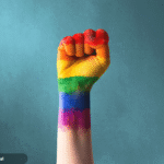Foto de um punho cerrado com as cores pintadas da bandeira LGBTQIA+.