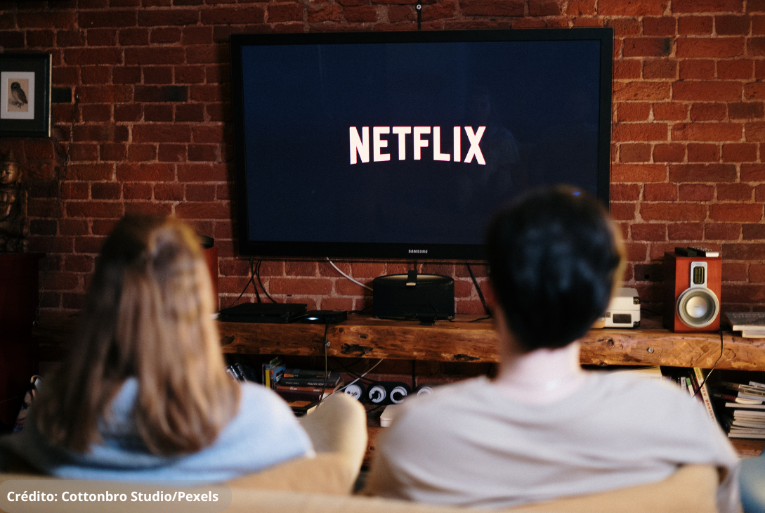 Imagem ilustrativa de um casal olhando para a TV com logotipo da Netflix.