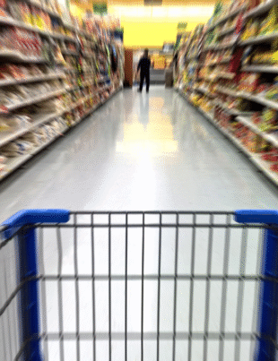 Foto ilustrativa do interior de um supermercado com prateleiras longas e repletas de produtos dos dois lados de um corredor. Em primeiro plano, vê-se um carrinho de compras, sugerindo a perspectiva de um consumidor empurrando-o pelo corredor.