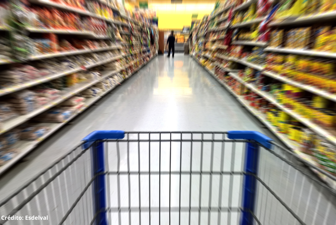 Foto ilustrativa do interior de um supermercado com prateleiras longas e repletas de produtos dos dois lados de um corredor. Em primeiro plano, vê-se um carrinho de compras, sugerindo a perspectiva de um consumidor empurrando-o pelo corredor.