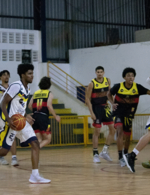 Na imagem, vemos um momento de ação durante uma partida de basquete entre as equipes do Basquete Avaré (uniforme branco com detalhes em azul e amarelo) e Sorocaba Basquete (uniforme preto com detalhes em vermelho e amarelo). Um dos jogadores do Basquete Avaré conduz a bola pela quadra, com olhar atento e postura ofensiva.
